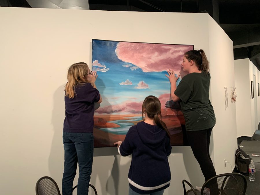 Student artists (from left) Eren Hall-Williams, Taylor Bauman, and Ann McSparrin hang a painting on the wall of USAOs Nesbitt Gallery as part of the Bachelor of Fine Arts Show that will be on display through Dec. 7.